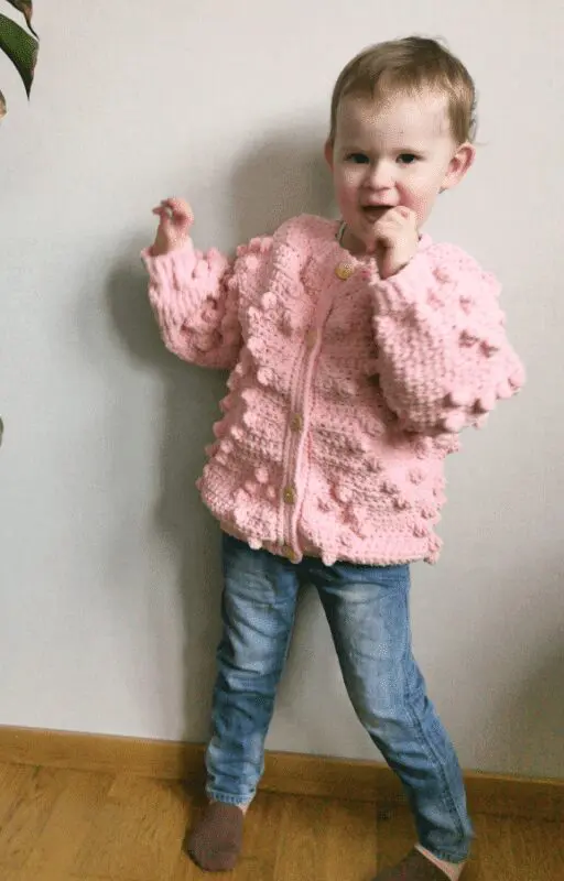 Toddler wearing a pink crochet heart cardigan with bobble stitches, shown from front and back. Includes close-up of stitch detail and SweCraftCorner branding. Free crochet pattern featured for babies and toddlers.