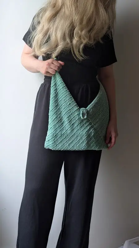 Crochet Bag - Free Pattern - Make it in 1 weekend Woman holding a handmade mint-green crochet bag with textured stitches and a decorative clasp, styled with a black jumpsuit. The crochet bag is made using the free bag crochet pattern in the article