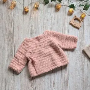 baby wrap cardigan crochet pattern in soft pink yarn, long sleeves, flat lay on light wood background