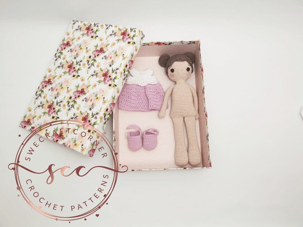 A floral gift box lies open, revealing a handmade amigurumi doll in a pink and white dress, with matching pink shoes neatly arranged inside. The charming crochet toy set is beautifully displayed on a plain white surface.