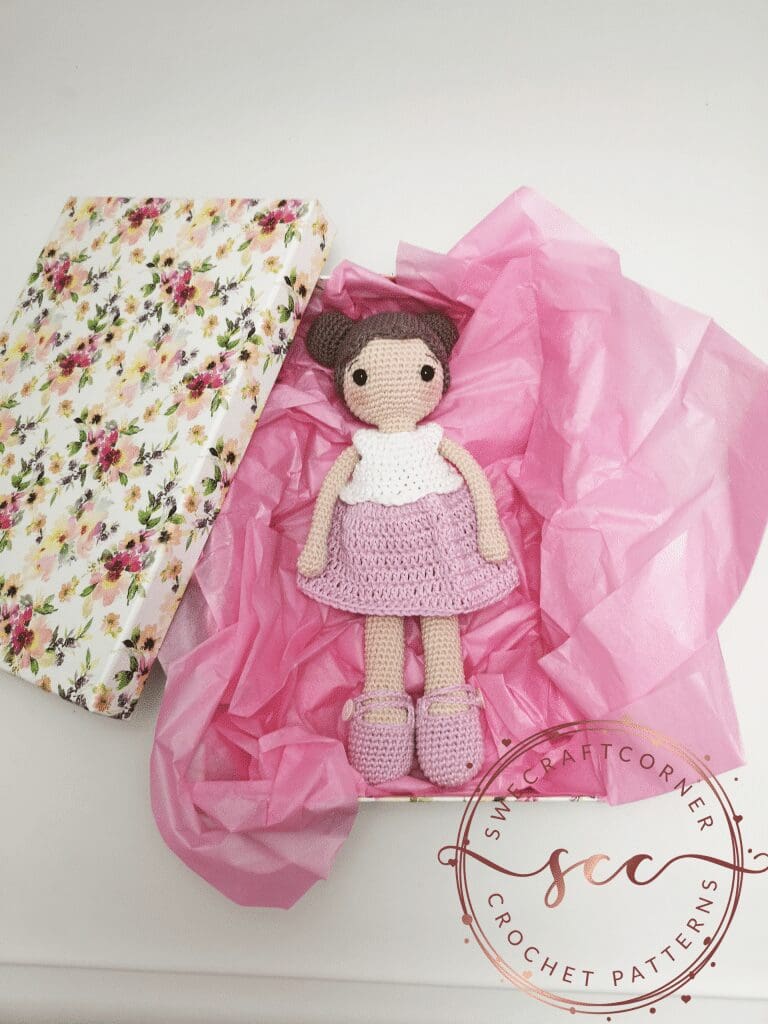 An amigurumi doll with brown hair, a white top, pink skirt, and matching shoes lies on pink tissue paper in a floral gift box. The lid is beside her, and a crochet patterns logo is visible in the corner.