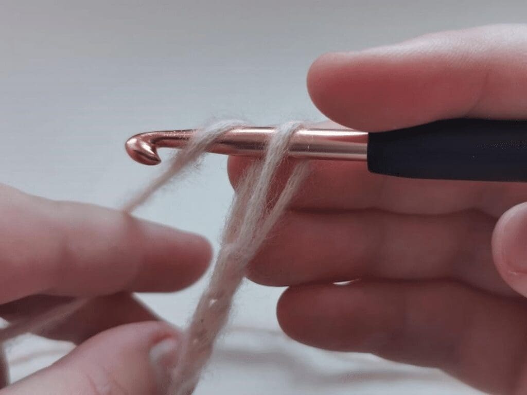 Close-up of hands holding a crochet hook and beige yarn, preparing to crochet. The yarn is looped over the hook as fingers adjust the strands, ready to demonstrate how to crochet the jasmine stitch.