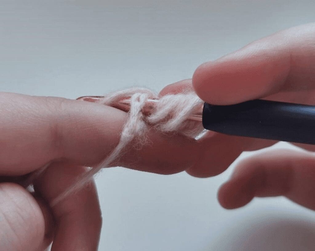 Close-up of hands holding a crochet hook and light-colored yarn, with the yarn looped around the hook and fingers, showing the first steps of how to crochet the jasmine stitch.