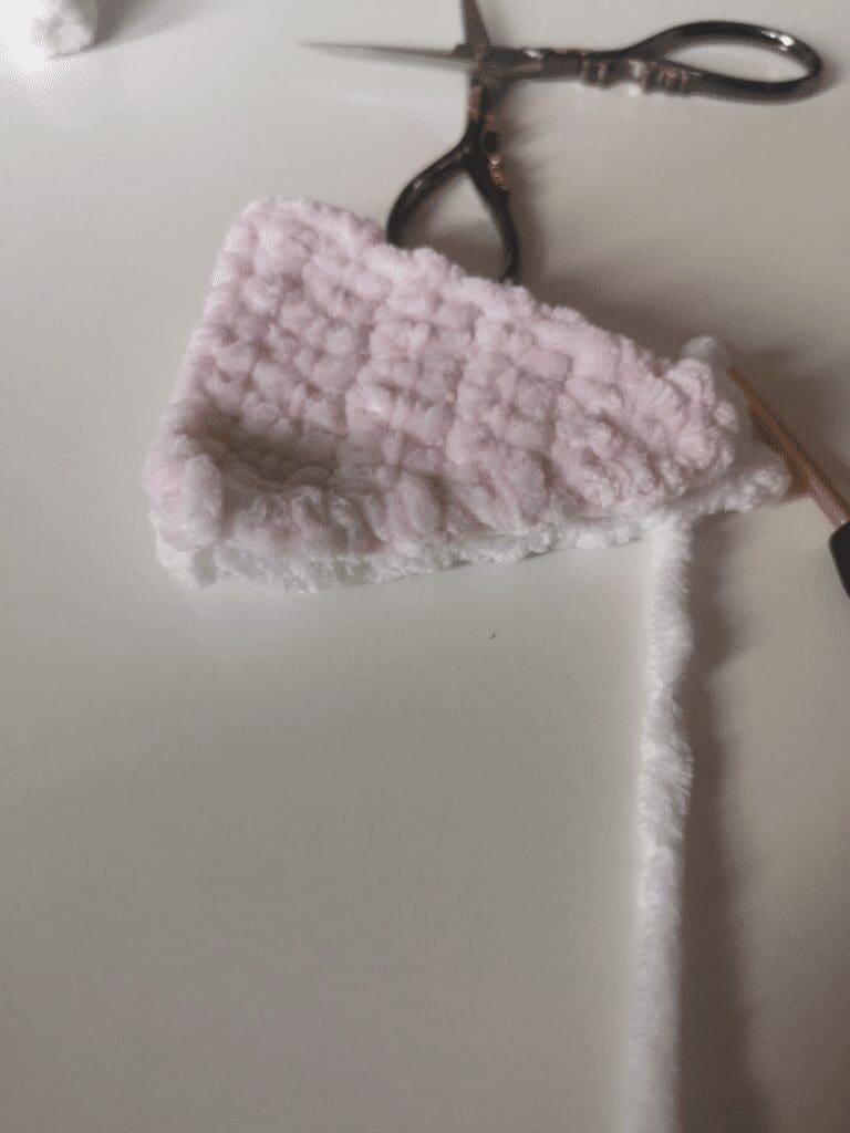 A partially crocheted piece of soft, light pink and white textured yarn—soon to become a crochet cat plushie—rests on a white surface beside a pair of scissors and a crochet hook.