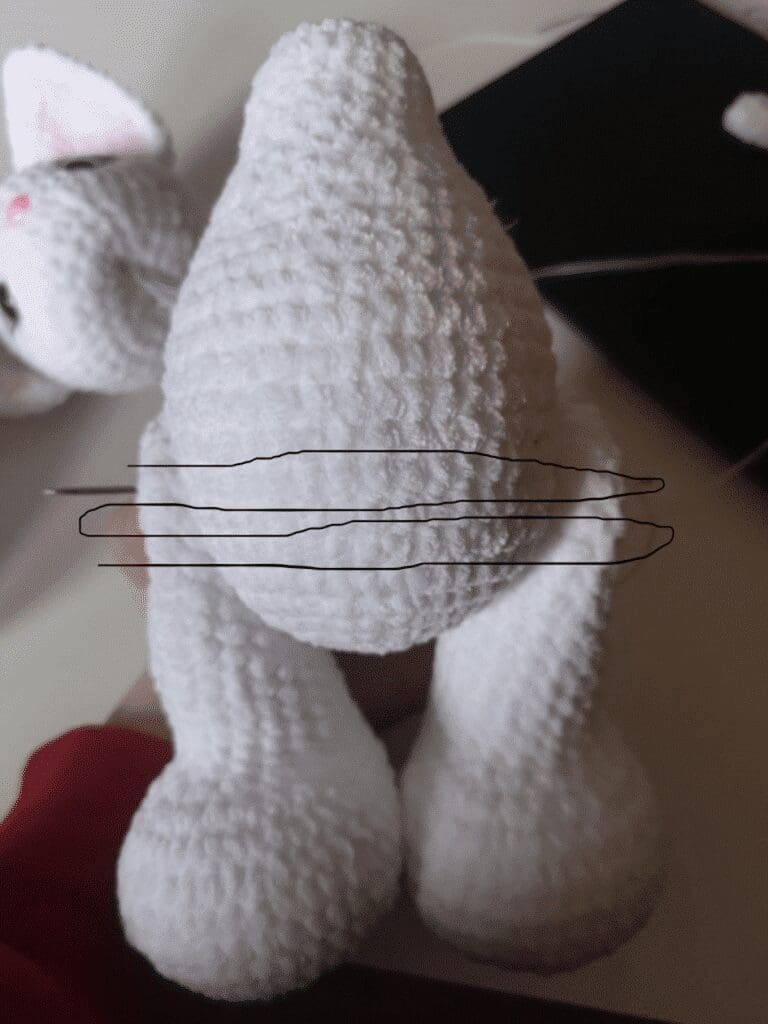 A crocheted white stuffed animal body with legs, destined to become a crochet cat plushie, is held upright. A partially attached ear and unfinished crochet parts are visible in the background.
