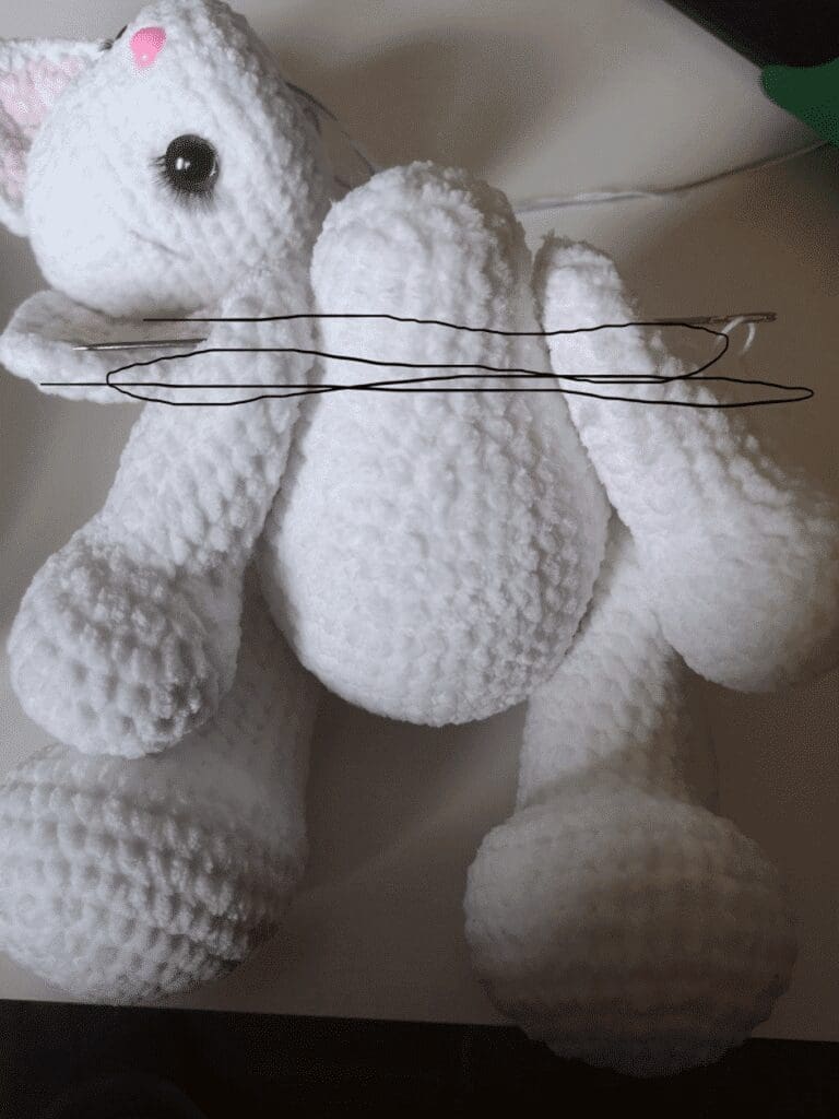 A white textured stuffed animal resembling a bunny with a pink nose and black eyes lies on a surface, similar in style to a crochet cat plushie. Two black lines are drawn across its body near the arms.
