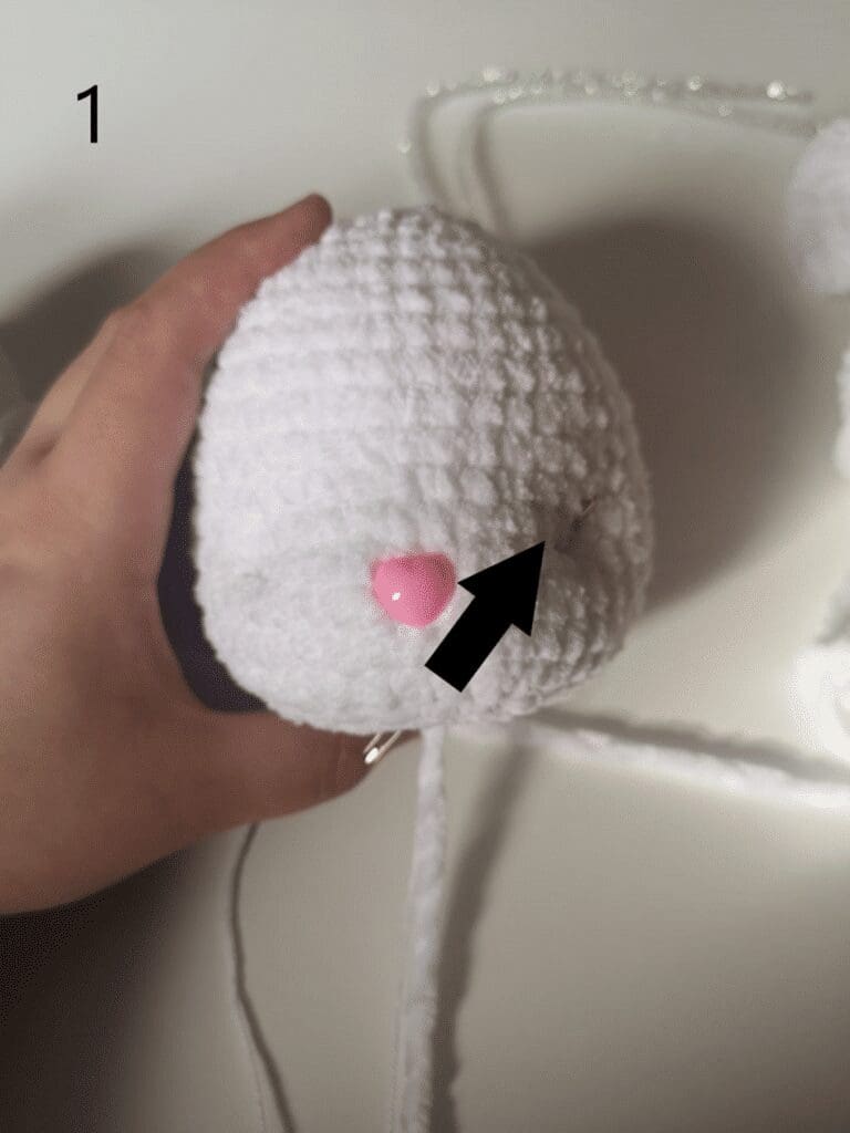 A hand holds a white crocheted oval shape, part of a crochet cat plushie. A pink pin is inserted into the crochet, with a black arrow pointing to it. The number 1 appears in the top left corner.