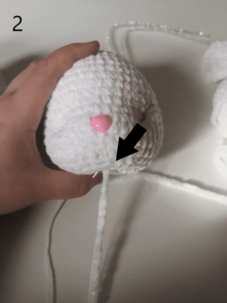 A hand holds a white crochet cat plushie ball with a pink nose attached. A needle is inserted into the ball, and a black arrow points to a white yarn strand coming out of the bottom. The number 2 is in the top left corner.
