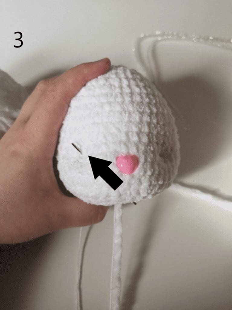 A hand holds a white, textured yarn ball with a needle inserted—perfect for crafting a crochet cat plushie. A black arrow points to a pink, heart-shaped bead attached to the yarn. The number 3 appears in the top left corner.