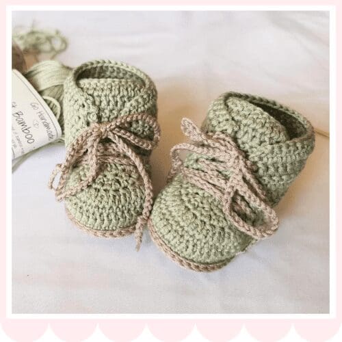 A pair of hand-crocheted green baby booties with beige laces, perfect for complementing other baby crochet clothes, are displayed on a white surface with a skein of yarn in the background.