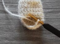 A crochet hook with a black handle is working with white yarn on a small crocheted piece for baby crochet clothes, placed on a gray wooden surface.