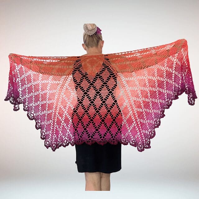 A person with light hair in a bun wears a black dress and holds out a large, lacy, crochet shawl in shades of orange, pink, and purple, displaying its intricate pattern against a plain background.