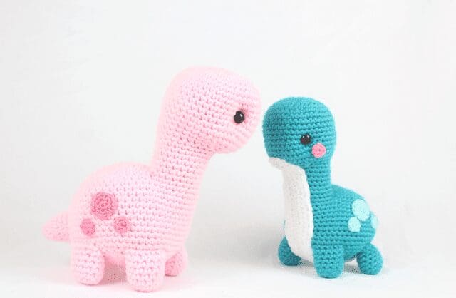 Two crochet dinosaur toys, one pink with darker pink spots and the other teal with a white belly and blue spots, face each other against a plain white background.