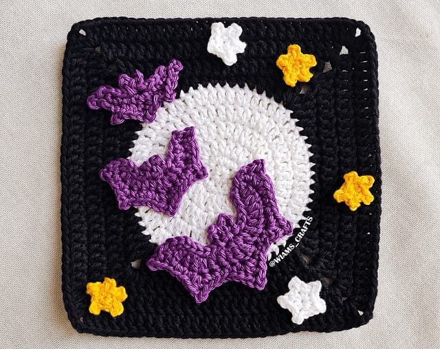 A crochet square with a white circle in the center, surrounded by a black border. Three purple bats and five yellow and white stars are attached, creating a spooky night scene.