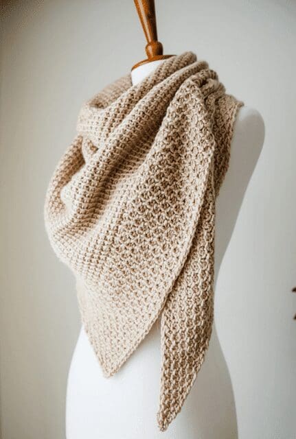 A beige, textured crochet shawl is draped over a white mannequin torso, displayed against a plain background. The shawl features a triangular shape and a detailed, waffle-like stitch pattern.