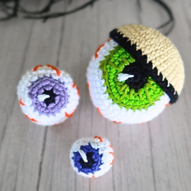 Three crochet eyeballs of different sizes and colors—green, purple, and blue—are displayed on a light wooden surface; the largest has a tan eyelid partially covering it.