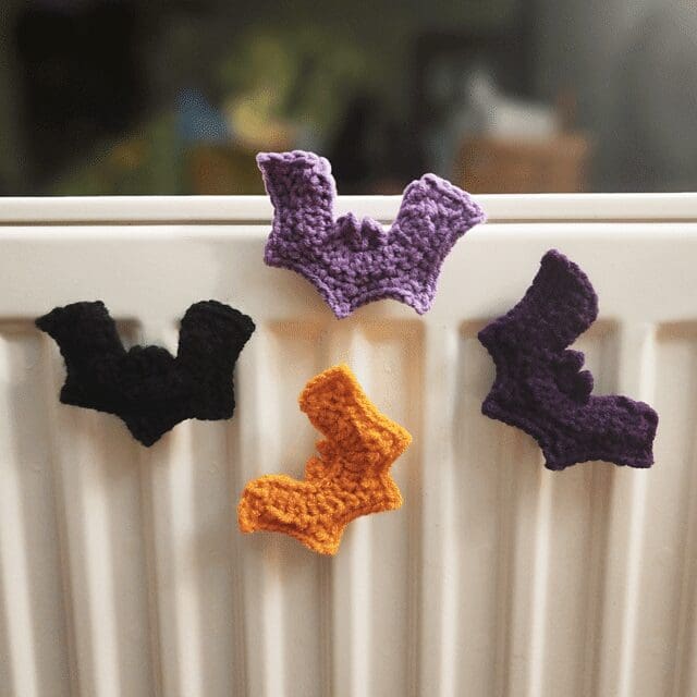 Four crochet bat shapes in black, purple, and orange are displayed on a white radiator. The bats are simple and whimsical, suggesting a Halloween or autumn theme. The background is out of focus.