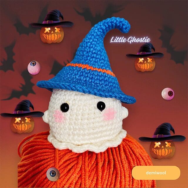 A crochet ghost wearing an orange sweater and a blue witch hat is surrounded by spooky cartoon pumpkins and eyeballs on a Halloween-themed background. Text reads Little Ghostie and demiwool.