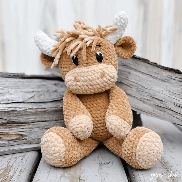 A plush, crochet cow toy with light brown and cream-colored yarn sits on wooden planks in front of weathered wood. The cow features white horns, black eyes, and a tuft of yarn hair on its head.