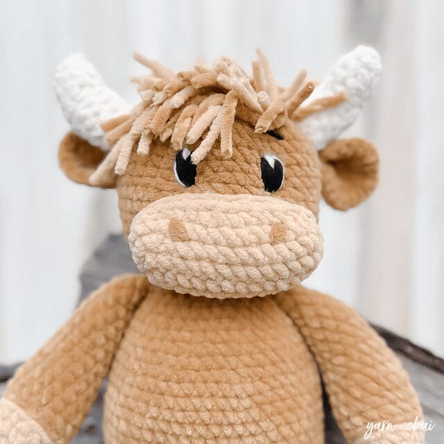 A handmade, crochet plush bull in light brown and beige with white horns and yarn hair, featuring large black eyes and a neutral expression, sits against a blurred background.