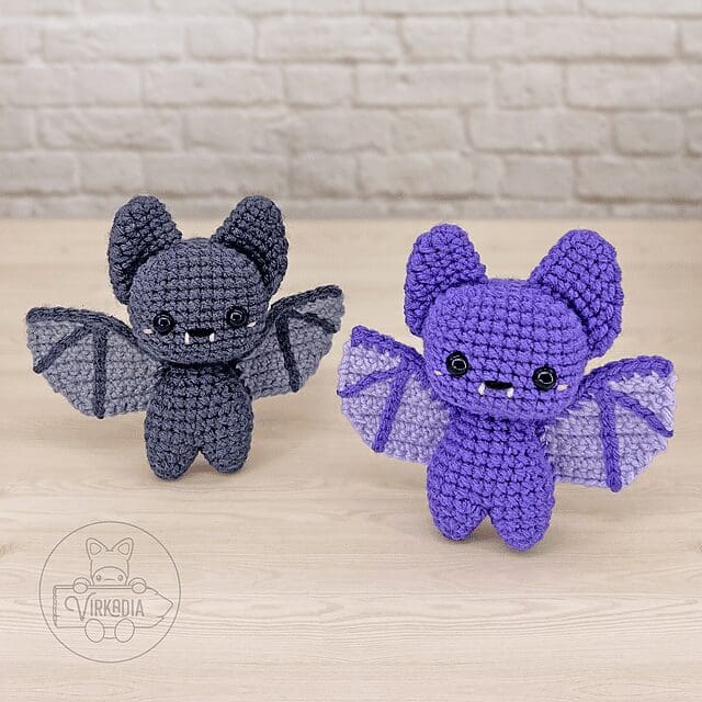 Two crochet bat dolls, one dark gray and one purple with light gray wings, stand on a light wooden surface against a white brick wall. Both feature big eyes, small fangs, and outstretched wings.