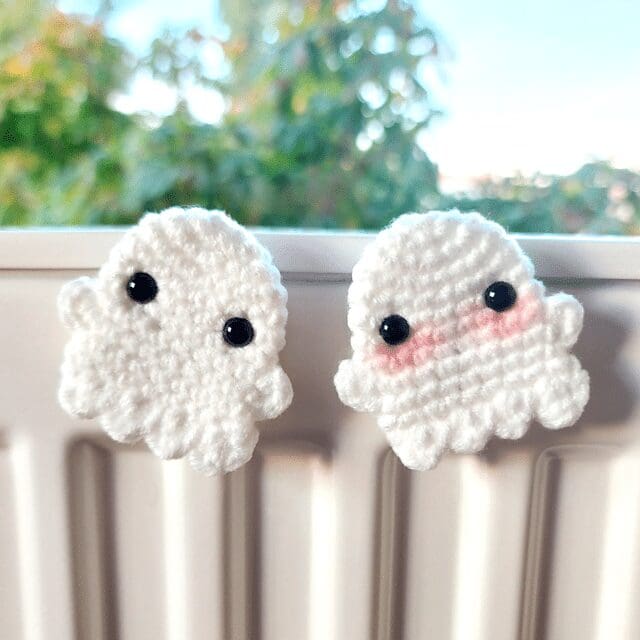 Two small crochet white ghost figures with black eyes sit side by side on a windowsill; one has pink cheeks. Green foliage is blurred in the background.