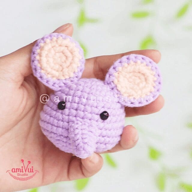 A hand holds a small crochet lavender mouse head with large round ears and black eyes. The mouse has a short snout, and the softly blurred background features green and white hues.