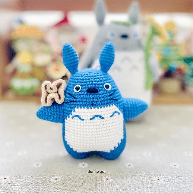 A crochet blue Totoro plush toy with a white belly and small beige flower stands on a light patterned surface. Blurred, colorful crochet figures are in the background.