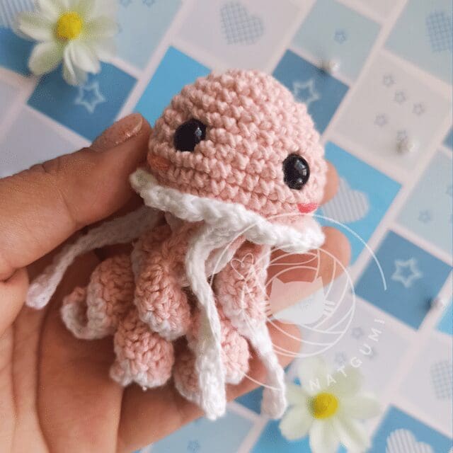 A hand holds a small, pink crochet jellyfish with big black eyes and white tentacles. The background features a blue and white patterned surface adorned with flowers and geometric shapes.