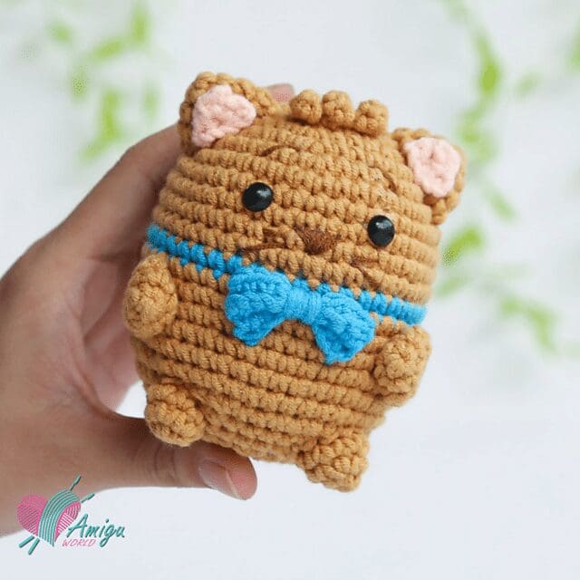 A hand holds a crochet brown cat plush with black eyes, a blue bow, and pink inner ears. The background is white with green, blurred shapes. A logo in the bottom left reads Amiga World.