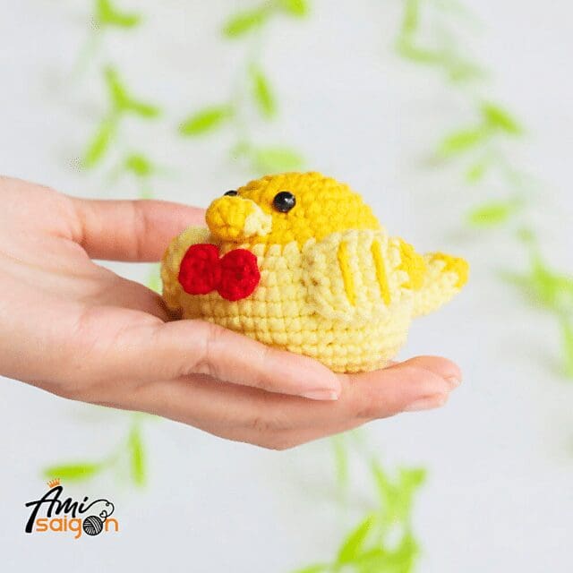 A hand holds a small, crochet yellow chick with a red bow tie. The background is white with green, blurred leaves, and the Ami Saigon logo is visible in the corner.