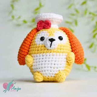 A crochet stuffed animal resembling a yellow and white dog with orange ears, black eyes, a white muzzle, a pink bow, and a chef’s hat sits upright against a blurred leafy background.
