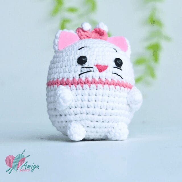 A crochet white cat plushie with a pink crown and collar, pink ears, and black embroidered eyes, nose, and mouth stands against a light background with blurred green foliage behind.