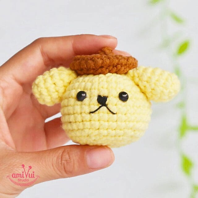 A hand holds a small, yellow, crochet bear head with round ears, black eyes, a simple mouth, and a brown crocheted beret. The blurred background shows hints of green.
