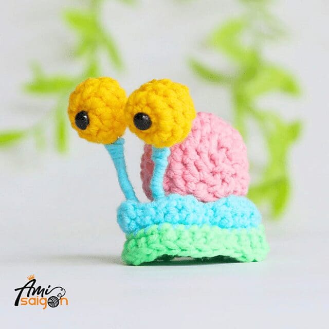 A crochet snail with a pink shell, blue and green body, and large yellow eyes on blue stalks sits on a white surface, with green leaves blurred in the background. The Ami Saigon logo is in the bottom left corner.