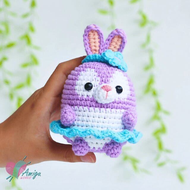 A hand holds a small, crochet purple bunny with a blue skirt, blue hat, and pink inner ears. The bunny has black eyes, a white snout, and is set against a blurred leafy background.