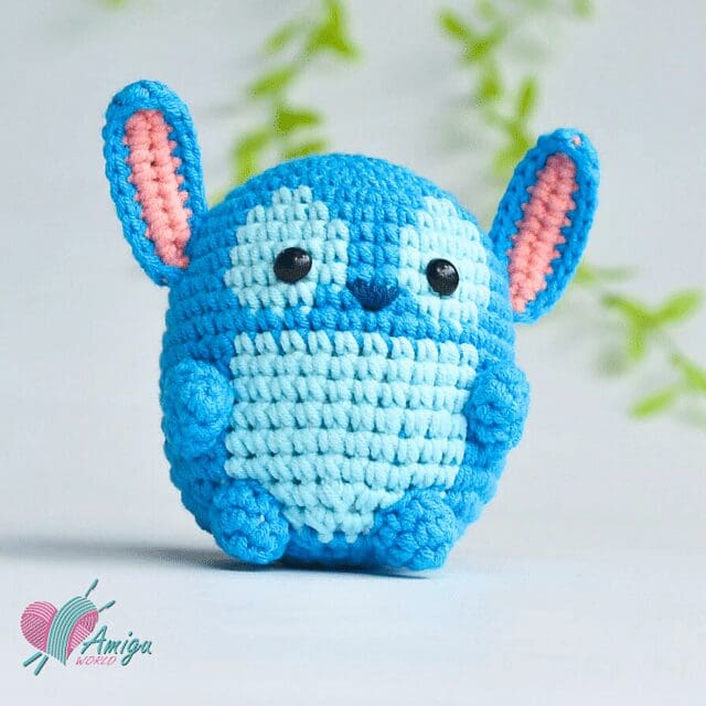 A crochet blue plush toy with large round eyes, a small smile, and big ears with pink inner details. It has a light blue belly and sits upright on a white surface, with blurred green foliage in the background.