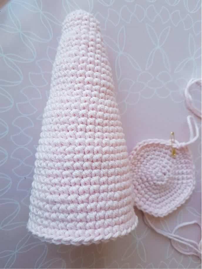A crocheted pale pink cone, part of a stacking toy crochet set, stands upright next to a flat crocheted pink circle with a yarn needle and yarn attached, both resting on a patterned surface.