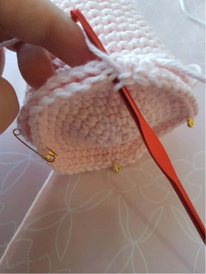 A close-up of a hand crocheting a stacking toy crochet piece with light pink yarn, using a red crochet hook. The cylindrical crochet work features stitch markers and a safety pin attached to the edges.