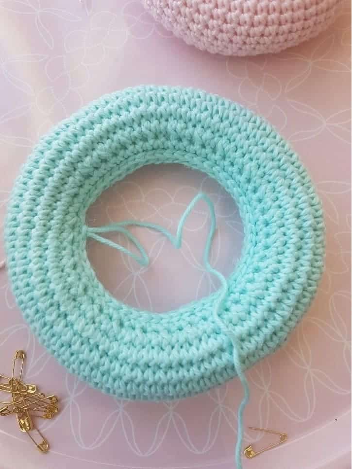 A light blue crocheted ring from a stacking toy crochet set, with a loose yarn tail, rests on a pale surface with a subtle floral pattern. Several gold safety pins are scattered nearby.