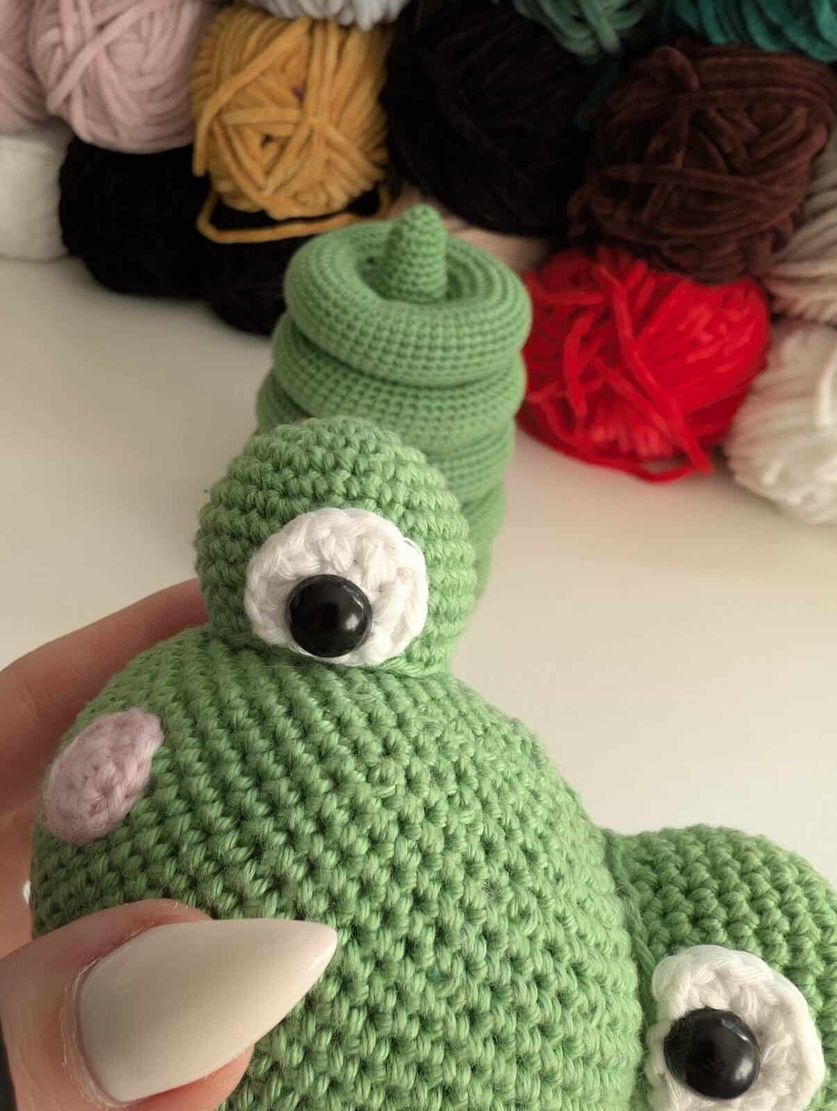 A close-up of a hand holding a crocheted green frog doll, with black and white eyes. In the background, colorful yarn balls and another stacking toy crochet piece add charm to the cozy scene.