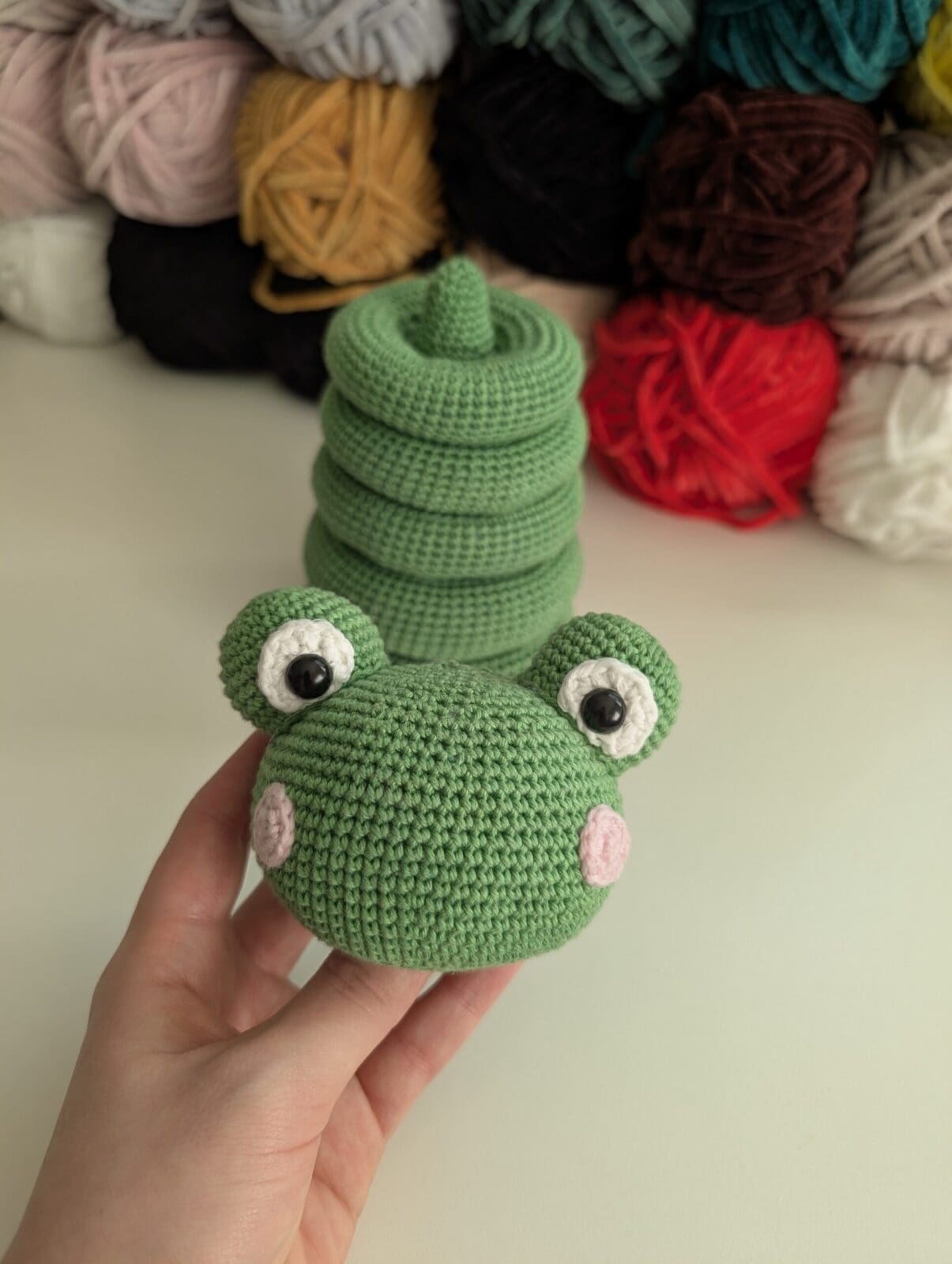 A hand holds a crocheted green frog toy with big eyes and pink cheeks, crafted in the stacking toy crochet style. Behind it, several skeins of yarn in various colors are arranged on a white surface.