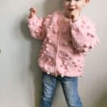 Toddler wearing a pink crochet heart cardigan with heart-shaped bobble stitches, posing playfully.