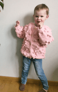 Toddler wearing a pink crochet heart cardigan with bobble stitches, shown from front and back. Includes close-up of stitch detail and SweCraftCorner branding. Free crochet pattern featured for babies and toddlers.