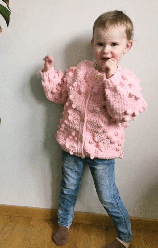 Toddler wearing a pink crochet heart cardigan with bobble stitches, shown from front and back. Includes close-up of stitch detail and SweCraftCorner branding. Free crochet pattern featured for babies and toddlers.