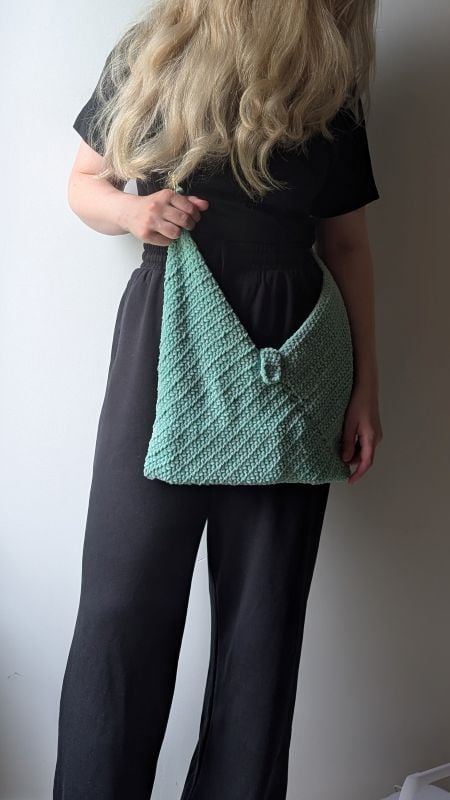 Woman holding a handmade mint-green crochet bag with textured stitches and a decorative clasp, styled with a black jumpsuit. The crochet bag is made using the free bag crochet pattern in the article