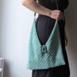 Woman holding a handmade mint-green crochet bag with textured stitches and a decorative clasp, styled with a black jumpsuit. The crochet bag is made using the free bag crochet pattern in the article