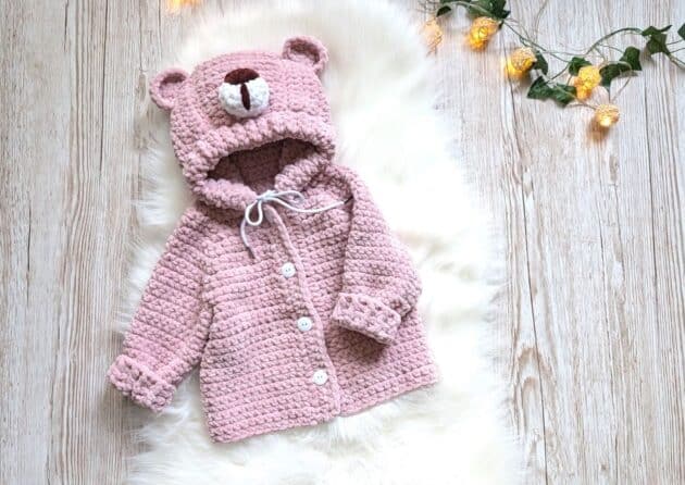 Crochet baby hoodie with bear ears and buttons made in soft pink chenille yarn, laying on a wooden background – Bearly Adorable Jacket by Swecraftcorner.