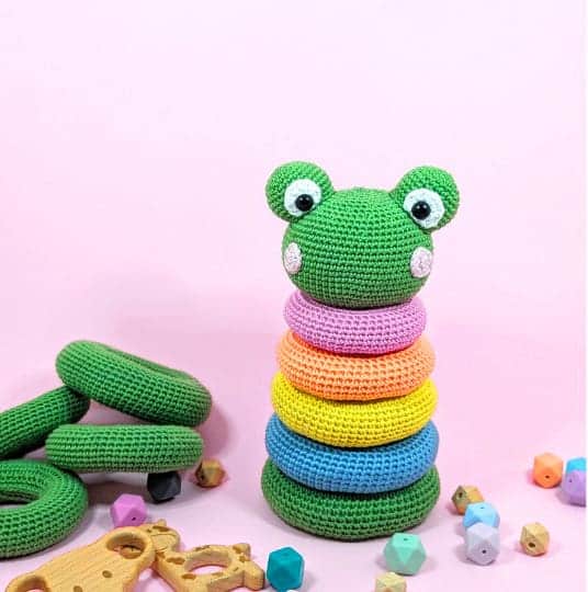 Frog stacking toy crochet pattern shown as a crocheted green frog head with colorful rings on a pink background