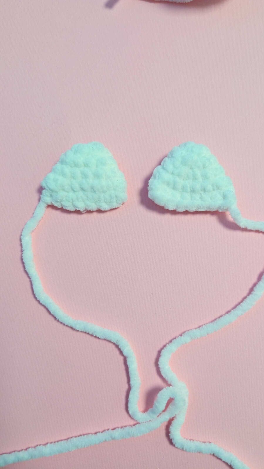 Two fluffy white pom-pom shapes connected by a fuzzy yarn string, resembling a bikini top, are laid out on a soft pink textured background—perfect inspiration if you're learning how to crochet.